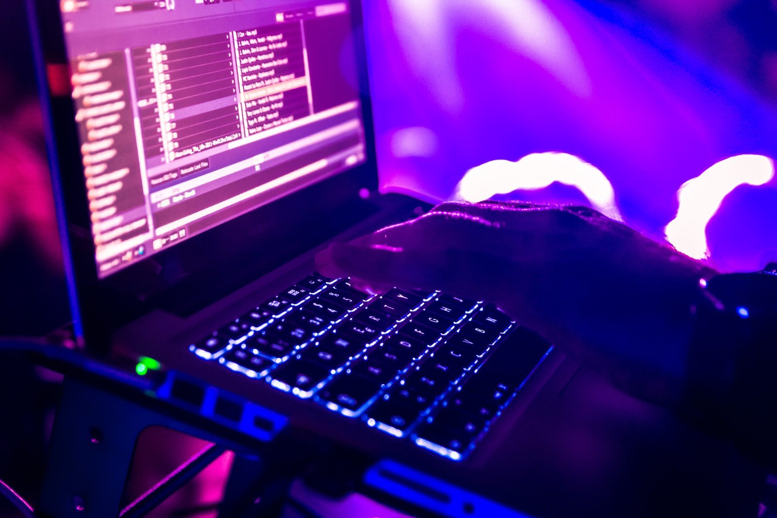 A person’s hand operates a laptop with illuminated keys, displaying audio software and surrounded by purple and blue stage lighting - a glimpse into the technology behind Our Services.